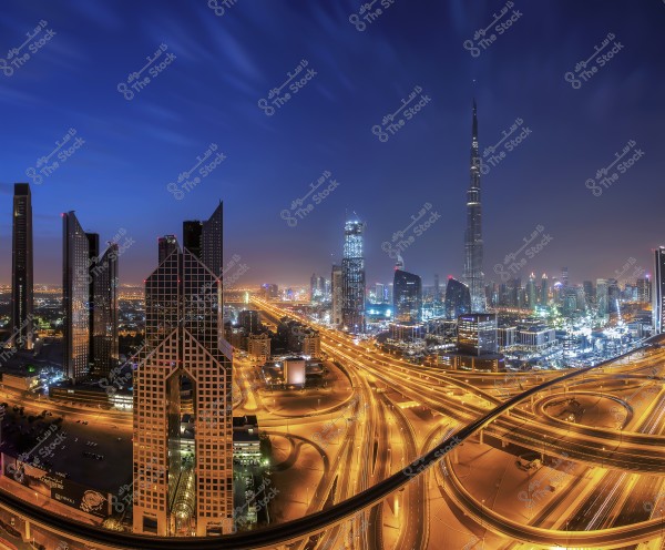 Nighttime image of Dubai city showcasing the illuminated skyscrapers, including the towering Burj Khalifa in the skyline. The intertwined highways form a glowing network while the light from the buildings reflects in the deep blue sky.
