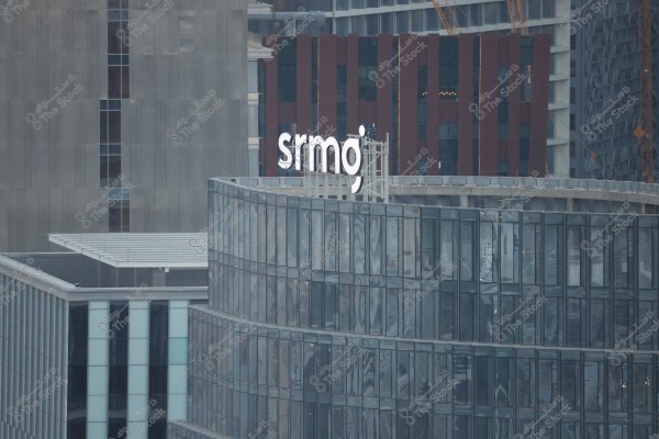 An image of a modern building with large glass facades. The building has a white sign with the name \"srmgo\" on it. In the background, there is another contemporary building with dark colors.
