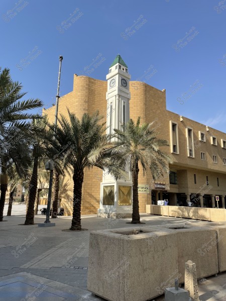 A picture of a clock tower located near a building in a modern architectural style, with brown brick walls. The tower appears with a green roof and around it palms planted on the site. The sky is clear and blue, indicating a sunny atmosphere. The building is large and casts shadows on the surrounding area.