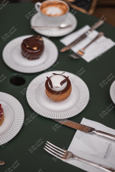 صورة تعرض قطع حلوى مختلفة مرتبة بشكل أنيق على طاولة خضراء, بجانبها فنجان قهوة وأدوات مائدة فضية.