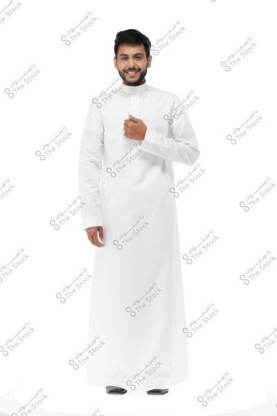 A photo of a man standing and smiling in a white studio. The man is wearing a white thobe, a traditional garment common in Gulf countries like Saudi Arabia. He is holding one side of the thobe with his right hand and appears confident and at ease.