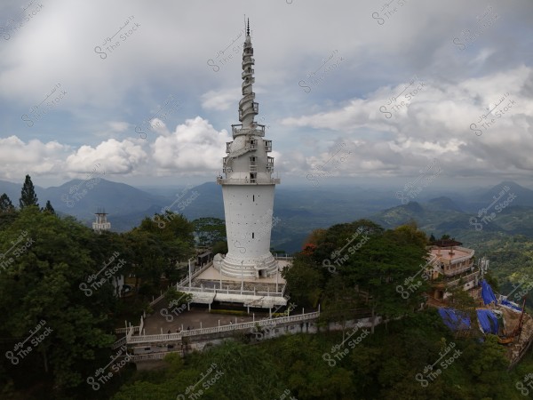 An image of a tall white tower with a spiral design, surrounded by a scenic landscape. The tower is situated on a hilltop surrounded by green trees, with a mountain range visible in the distance under a partly cloudy sky. Nearby buildings and trees add a tranquil feel to the scene.