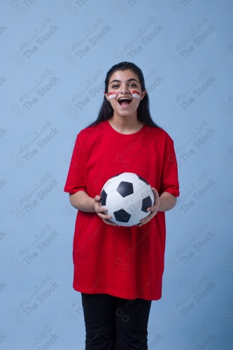 Portrait of a Saudi woman wearing a red T-shirt cheering the football team on a blue background and showing expressions of joy and enthusiasm, World Cup.