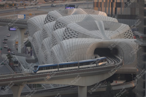مشهد لمحطة قطار بتصميم معماري حديث ومبهر في مدينة الرياض بالمملكة العربية السعودية. يظهر القطار على مسار مرتفع في المقدمة وهو يدخل المحطة التي تتميز بتصميم خارجي ديناميكي ومغطى بأشكال هندسية معقدة باللون الأبيض. المشهد يشمل بعض السيارات على الطرق القريبة من المحطة والبنية التحتية العصرية المحيطة بها.