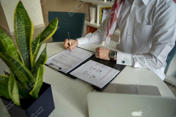 An image of a man wearing a white thobe and a red agal, sitting at a desk and filling out forms or documents. He has a watch on his wrist, with a laptop and a plant in the background. The setting is a neat office environment with a chair and a bookshelf.