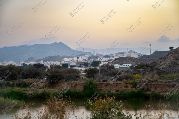 منظر طبيعي يضم مياه بركة صغيرة وأشجار متناثرة في المقدمة مع مدينة صغيرة في الخلفية. تظهر المباني الحديثة والبيضاء المدينة تندمج مع الطبيعة المحيطة. توجد جبال زرقاء خفيفة في الأفق تحت السماء الصافية وقت الغروب.