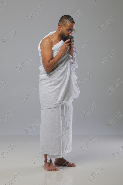 Image of a man wearing a white Ihram, the traditional garment worn by Muslims during the Hajj or Umrah pilgrimage. The man is standing on a gray background, barefoot, and holding a piece of cloth or a small container close to his face with a focused expression.