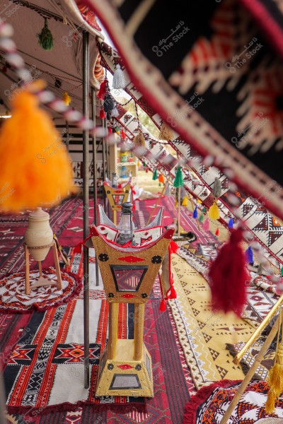 The image shows the inside of a traditional tent adorned with various Arabic textiles. The floor is covered with patterned rugs in red, black, and white colors. There are decorated covers and tassels in yellow, red, and green hues. In the center of the tent, there is a long decorative stand, carrying a water bottle, featuring traditional artistic details.
