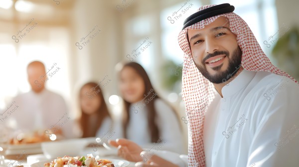 صورة لرجل يرتدي الثوب السعودي والغترة البيضاء والعقال الأسود, جالسًا مع عائلته حول طاولة طعام مزينة بأطباق متنوعة في مكان مضيء. يظهر في الخلفية أفراد عائلته يجلسون بجانبه في جو من الود والسعادة.