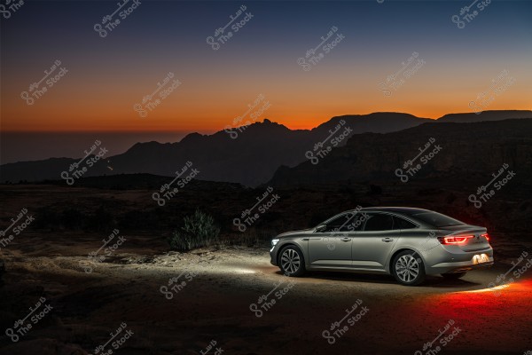 A silver car parked in a mountainous area at sunset. The sky is multicolored with shades of orange and dark blue, and the mountains appear as silhouettes in the background. The car\'s headlights and taillights are on, casting light onto the surrounding ground.