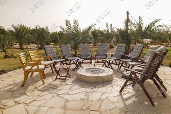 The image shows an outdoor seating area in a garden with wooden chairs featuring gray cushions, arranged in a circle around a fire pit. There are several small tables between the chairs. The surroundings include palm trees and greenery. The location has a stone floor and plants scattered in the distance under a clear sky.