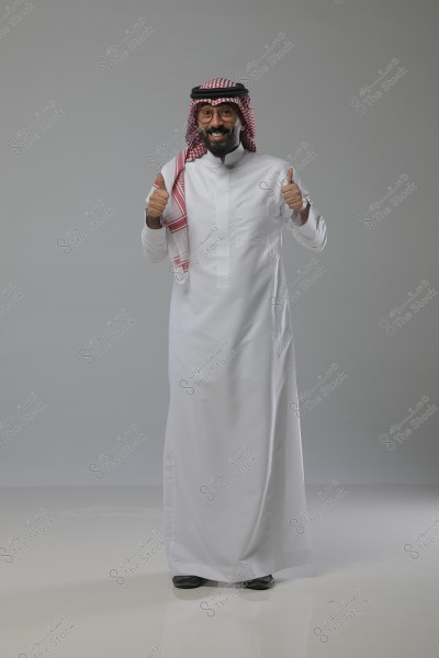 A portrait of a man standing in a studio, wearing a traditional Saudi white thobe with a red and white checkered shemagh and an agal. He is smiling and giving a thumbs-up with both hands. The background is plain gray.