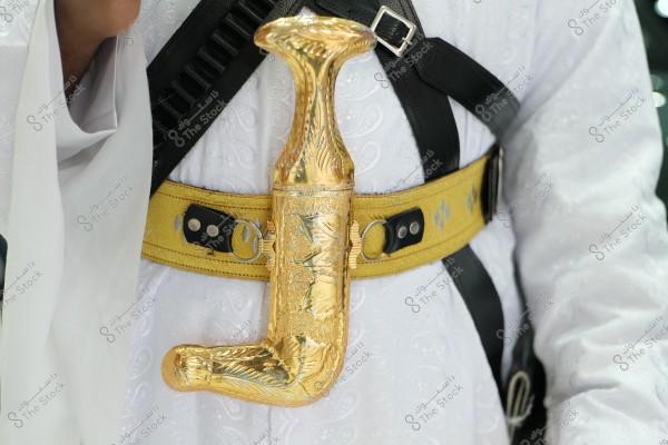 An image showing a person wearing traditional attire, featuring an ornate dagger with a golden handle placed on a golden belt with black details. The dagger is part of traditional clothing in some Arabian countries, such as Yemen and Saudi Arabia. The clothing is made of patterned white fabric.