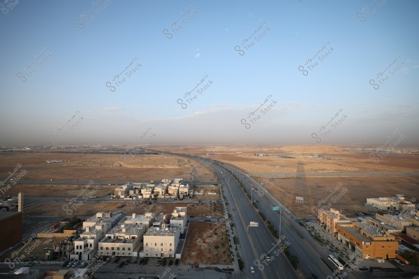 Aerial view of a semi-desert area with a highway dividing the region into two halves, surrounded by modern residential buildings with simple architectural designs. The dry, brown land stretches into the horizon under a partly cloudy blue sky. A few cars are visible on the road, and the sunlight casts long shadows on the ground.