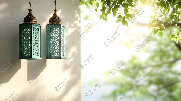 Two intricately designed green metal lanterns hanging against a light-colored wall, bathed in sunlight. The light filters through tree leaves in the background, creating a serene and natural atmosphere.