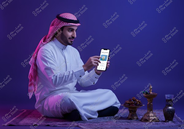 The image shows a man sitting on a decorative rug in a softly lit purple environment. He is wearing a white thobe and a white ghutra with a black agal, focusing on a mobile phone he is holding. Near him on the rug is an ornate table with plates of dates and a large bowl, as well as a traditional coffee pot.