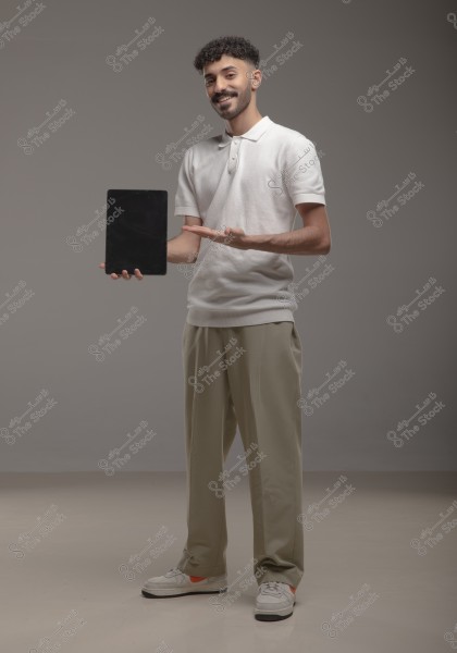 A portrait of a man standing in a studio wearing a white polo shirt and beige trousers. He holds a black tablet in one hand and gestures towards it with the other hand. He is smiling, and the background is a simple gray.