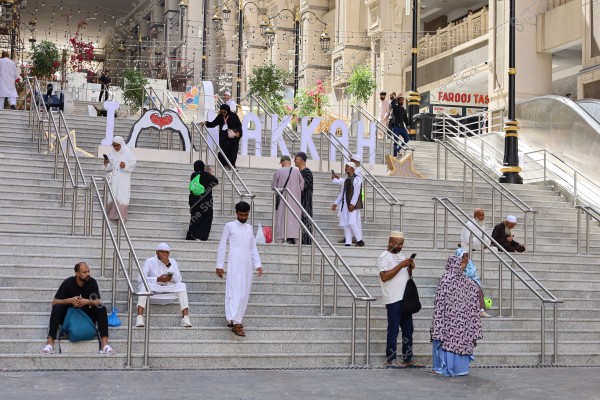 The image shows a group of people ascending and descending a large staircase leading to the entrance of a building in Mecca. There is a large sign that reads \"I ♥ MAKKAH,\" adorned with lights and plants on either side of the stairs. Individuals are dressed in various attire fitting the Islamic culture, such as white robes and abayas.