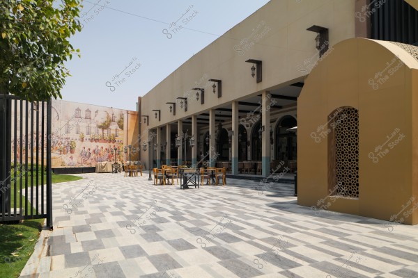 The image shows an outdoor courtyard of a modern building with a facade adorned with geometric patterns. Several tables and chairs are placed in the courtyard under an open roof supported by columns. The back wall is decorated with a mural depicting a traditional scene. There is also a small garden with trees on the left and a tiled flooring with a harmonious pattern.