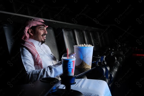 شاب سعودي يرتدي الزي السعودي التقليدي يجلس في صالة السينما ويظهر عليه السرور أثناء مشاهدة الفيلم، رجل سعودي يرتدي الزي السعودي التقليدي يجلس في صالة السينما ويظهر أمامه الفشار وأحد المشروبات على الطاولة