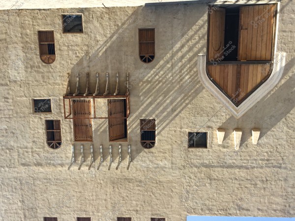 An image of a traditional building facade made of brick and plaster featuring wooden rectangular and arched windows. The windows of various sizes are closed with wooden latticework, and there is a large window protruding outward, showcasing wooden and decorative walls. The wall has sun rays casting shadows on its pale-colored surface, with a cool-colored roof visible in the background.