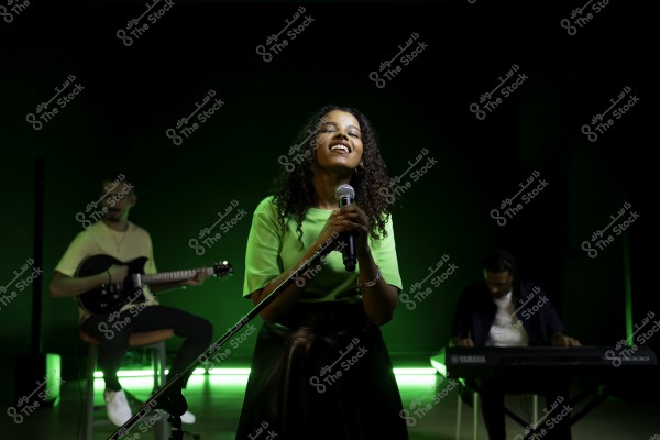 مغنية تقف على المسرح وتمسك ميكروفون, بينما يعزف موسيقيون على الجيتار والبيانو في الخلفية, والإضاءة خضراء.