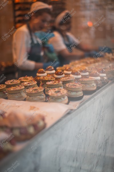 Display of a variety of decorated pastries inside a bakery with people working in the background.