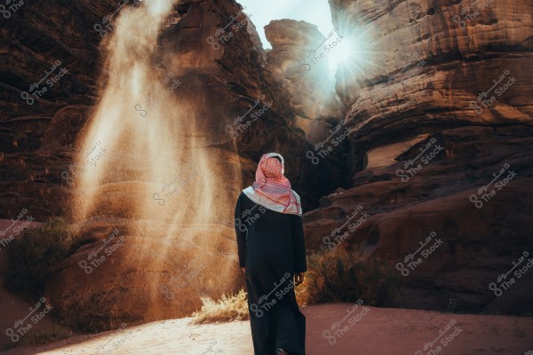 A person wearing a black robe and a red and white headscarf walks through a desert landscape with large rock formations illuminated by the sun from behind. The airborne dust creates a dramatic scene.