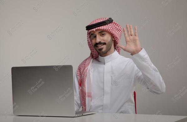 Portrait of a man sitting in front of a laptop, wearing traditional Saudi attire, including a white thobe, red and white checkered ghutra, and a black agal. He is raising his right hand in a friendly wave and smiling, seated on a regular chair in an indoor setting.