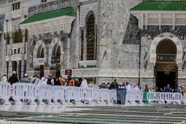 The image shows the facade of the Grand Mosque in Mecca, Saudi Arabia. Several worshippers and people dressed in white ihram and traditional clothing are walking near the large, ornate gates of the mosque. The facade is made of detailed marble with Islamic architectural features and adorned windows. The roof is covered with green tiles. There are white barriers and a decorated floor in the foreground.