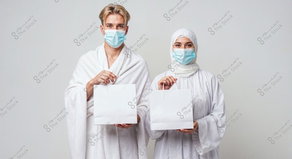 The image is a portrait of a man and a woman wearing white Ihram clothing for Umrah or Hajj, both wearing blue medical masks. The man wears a traditional Ihram robe, and the woman is wearing a white hijab. Each holds a white bag with one hand. The background is plain white.