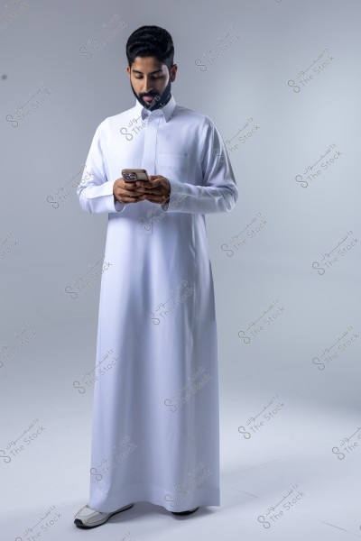 Image of a man wearing a white thobe and using a mobile phone. The man is standing in a studio with a gray background, and his features suggest he is from the Middle East. The overall impression reflects elegance and simplicity in traditional attire.