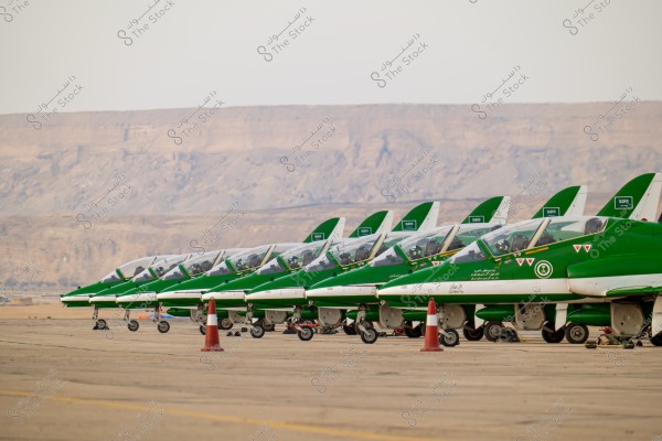 The image shows a lineup of green and white jet aircraft on an airport runway. The planes display markings indicating they are from Saudi Arabia, with mountainous terrain visible in the background. The aircraft bear the emblem and flag of Saudi Arabia.