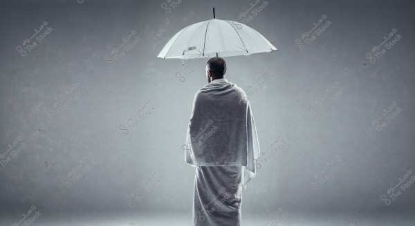 An image of a man wearing traditional white Ihram clothing, standing from the back and holding a large white umbrella. The background is a simple gray, emphasizing the details of the clothing and umbrella. This attire is commonly worn by Muslim pilgrims.
