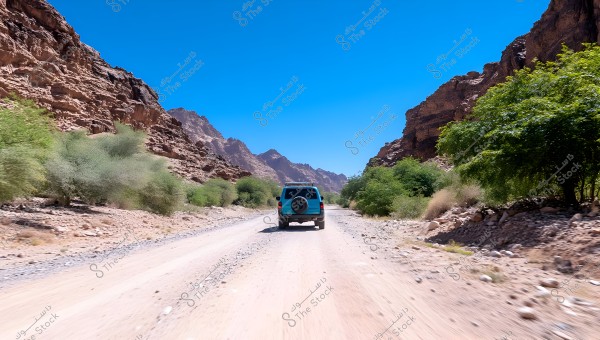 A blue car drives down a dirt road in a valley surrounded by rock formations and mountains. The road is flanked by green vegetation, and the sky is blue and clear in the background.