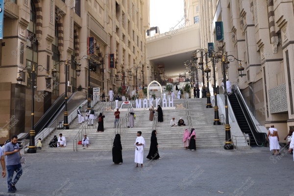 The image shows large steps located in Mecca with the words \"I love Makkah\" in white. People are wearing traditional Saudi clothing such as white thobes and black abayas, and some are performing Umrah. The surrounding buildings feature Islamic architectural designs. There are also people sitting and walking around the area.