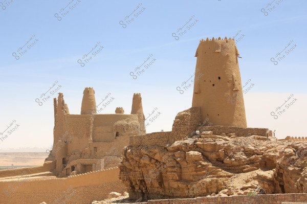 The image depicts an ancient fortress situated on a rocky hill on a sunny day. The fortress is constructed from mud and stone, featuring tall towers and sturdy walls. The fort\'s walls extend along the hill, enhancing its dramatic appearance. The sky is clear and blue, highlighting the details of the historical architecture.