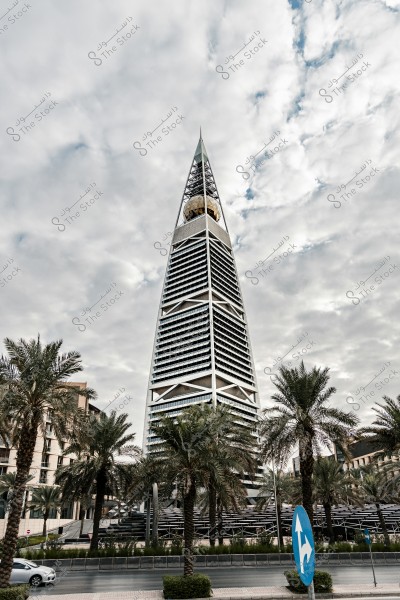 The Al Faisaliah Tower in Riyadh, Saudi Arabia, prominently displayed with its triangular shape and unique metallic structure topped with a golden glass orb. Palm trees and a traffic road surround the tower, with a cloudy sky in the background.