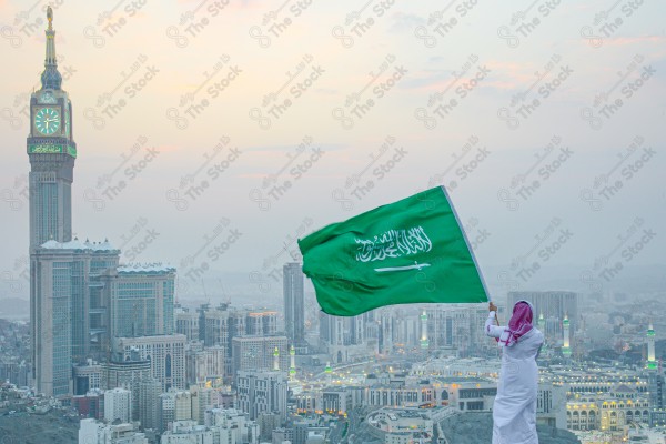 A snapshot of a Saudi man wearing the traditional Saudi dress, carrying the Saudi flag over a mountain in the Makkah Al-Mukarramah region, and the sky appears clear, the royal clock tower building in the Grand Mosque, buildings and landmarks, the Grand Mosque of Mecca.