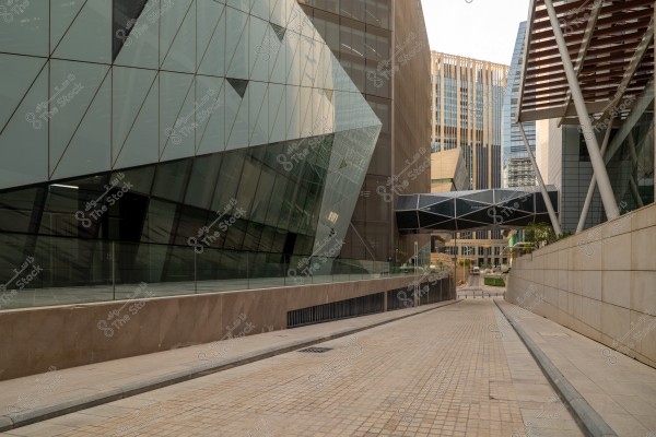 An image of an urban street surrounded by modern architectural buildings with sharp angles. The buildings are clad in glass and metal, with a glass pedestrian bridge connecting some of them above. The street is paved with stones and extends into the distance.
