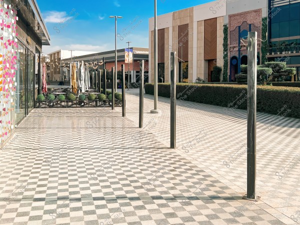 An image of an outdoor walkway at a shopping center, featuring a checkered black and white tiled floor. There are metal posts along the walkway and decorative plants on the sides. On the left side, there is an outdoor cafe with colorful umbrellas, and behind it, a building with modern architectural features.