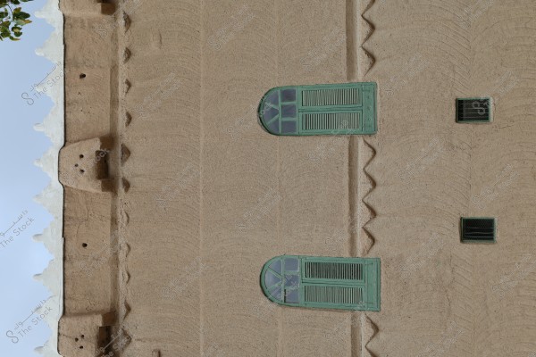 An image of a traditional mud-brick wall with green wooden windows in an Arabic style. The wall features traditional decorations and has a distinctive adobe structure. The top of the wall is adorned with prominent geometric elements.