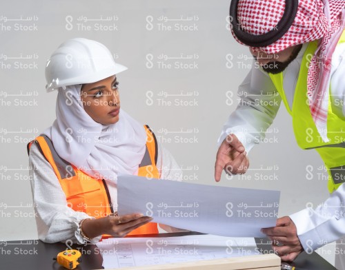 بورتريه مهندسة سعودية ترتدي الحجاب وسترة المهندسين وخوذة السلامة تمسك مع زميلها ورقة مخطط البناء وهي جالسة خلف الطاولة التي تظهر عليها أوراق لمخططات البناء وقطع خشب ومتر القياس بينما زميلها واقفًا يشير للمخطط