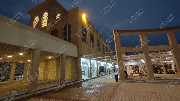 Nighttime image of a traditional market featuring a beige building with large windows and decorative arches illuminated by lights. Several open shops line the pavement, displaying clothes and other products at their entrances. An open ceiling with columns and dimly lit walkways is visible.