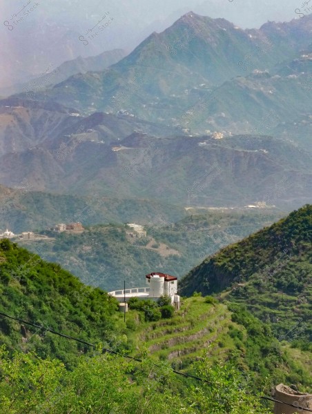 A scenic view of green terraced mountains in a rural setting. A white building with a red roof is situated on the hillside, surrounded by lush green trees. Scattered houses are visible in the background across the mountains under a clear blue sky.