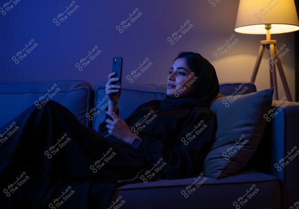 A photo of a woman sitting comfortably on a couch in a dimly lit room with blue lighting, holding a mobile phone. She is wearing a black abaya and a hijab. Beside her is a lit floor lamp, creating a calm and relaxed atmosphere.