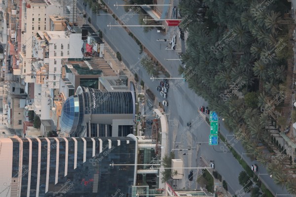 An aerial view of a city showing a main road intersection with multi-story buildings on the left and a cluster of trees on the right. The buildings have modern architectural designs, some with large glass windows. The road features traffic lights and directional signs in Arabic. Cars are orderly distributed along the streets.