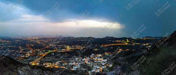 A nighttime cityscape with an aerial view of twinkling lights illuminating buildings and streets. Dark clouds cover the sky with faint light on the horizon indicating either sunset or sunrise. Winding roads stretch through the city, and surrounding hills add a scenic dimension to the view.