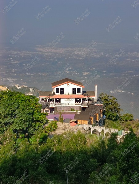 The image shows a large multi-story building located on top of a plateau, surrounded by lush greenery. The building features a spacious terrace on the upper floor with a sign that reads \"Restaurant.\" Numerous trees and dense vegetation surround the building, offering a serene natural view. The horizon extends in the background, covered by the shadows of hills and distant buildings under a clear blue sky.