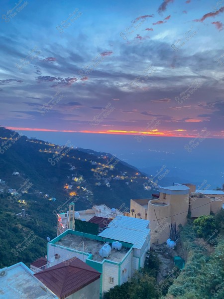 A scenic view of mountains with sunset color gradients, featuring houses on the mountain slopes and city lights in the horizon.